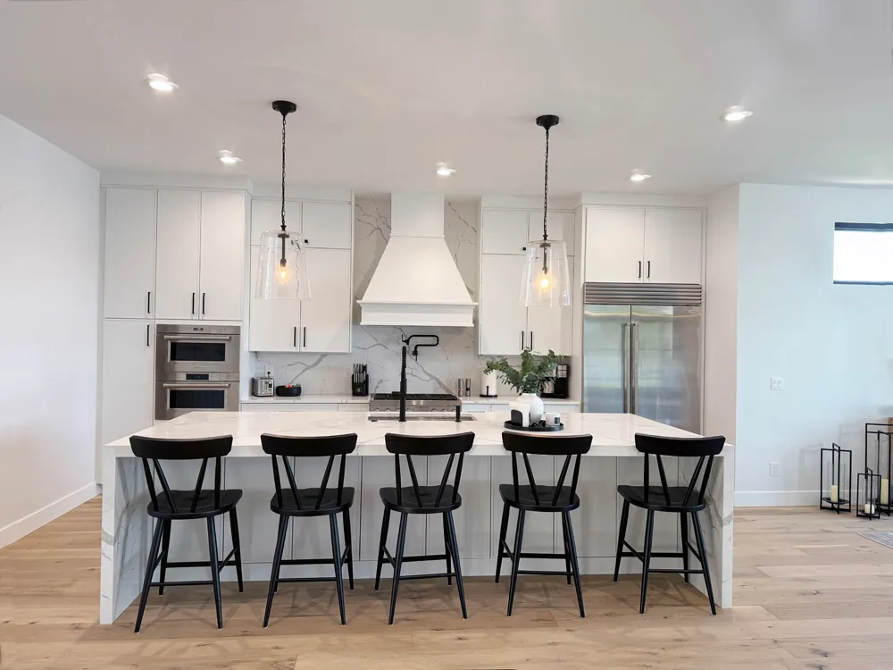 White kitchen with long waterfall island, five black chairs and white range hood