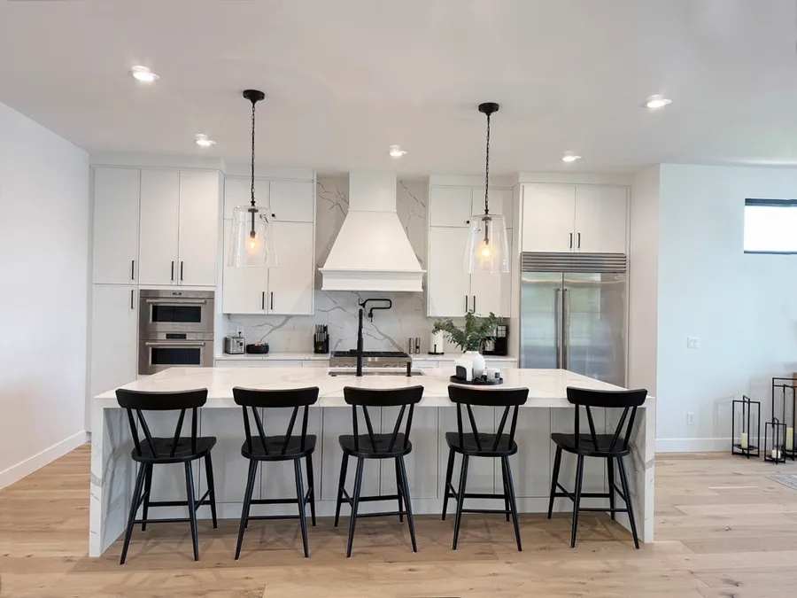 White kitchen with long waterfall island, five black chairs and white range hood