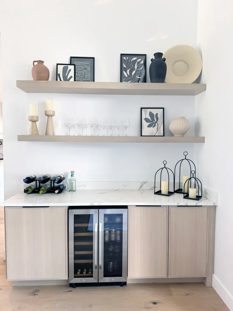 Dry bar with light wood cabinets, wine cooler and floating shelves of decor