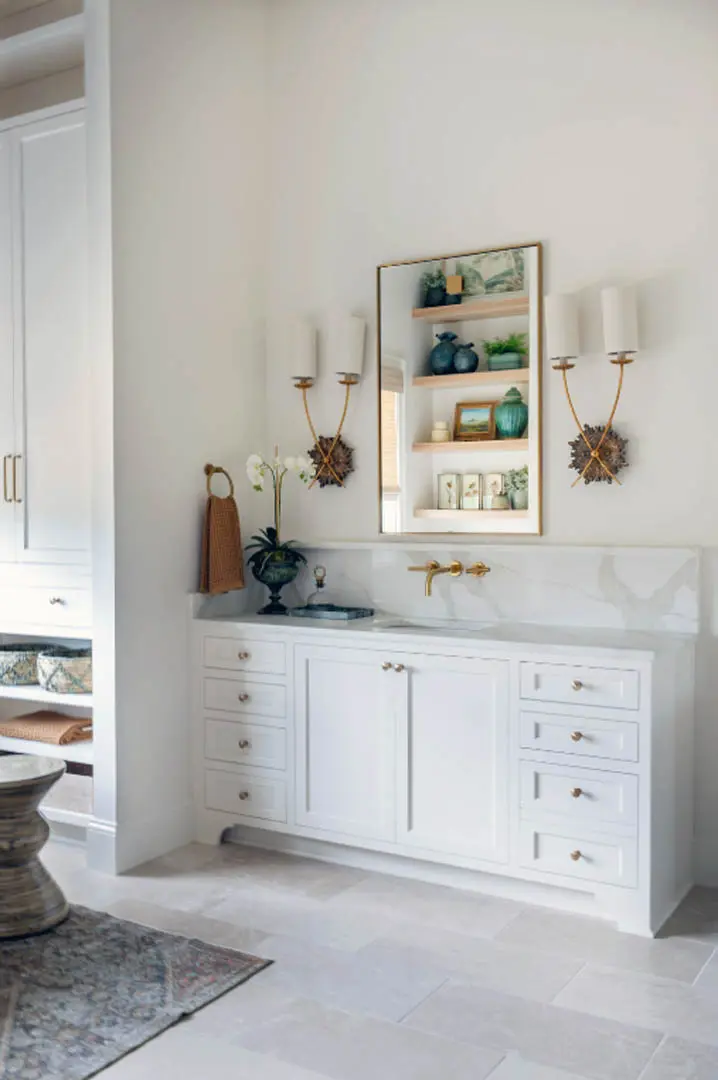 White bathroom vanity with marble backsplash, brass wall faucet and candle sconces
