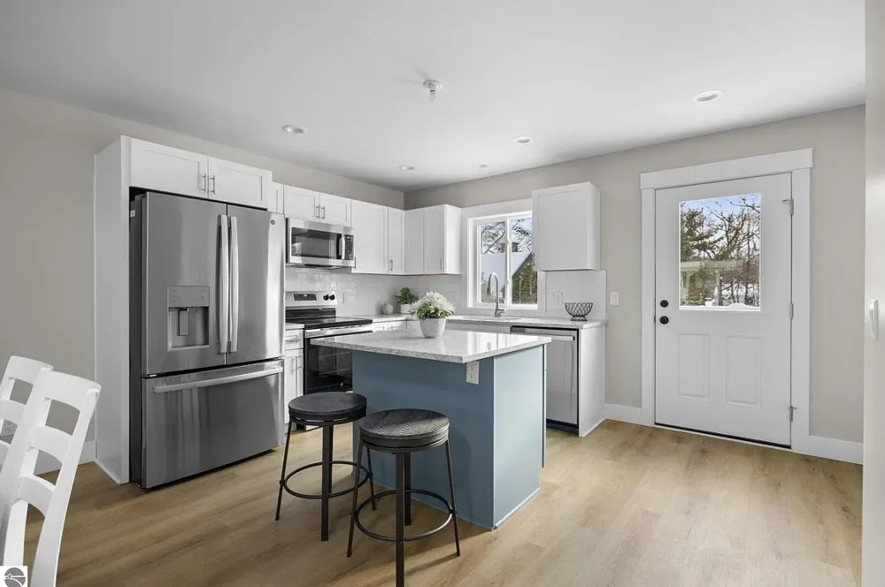 Small white kitchen with blue island, stainless appliances, two stools and a back door
