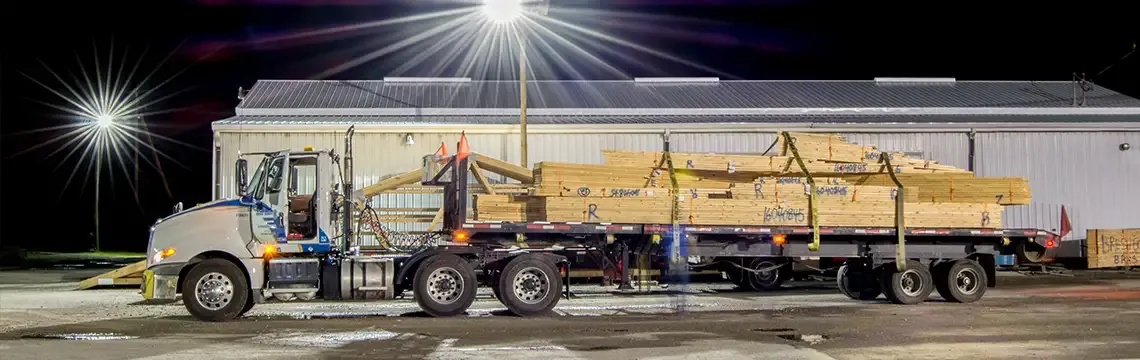 Flatbed truck loaded with wall panels and trusses outside a plant at night