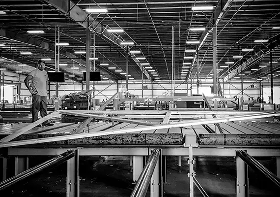 Black and white view of a worker at a long truss assembly table in a plant