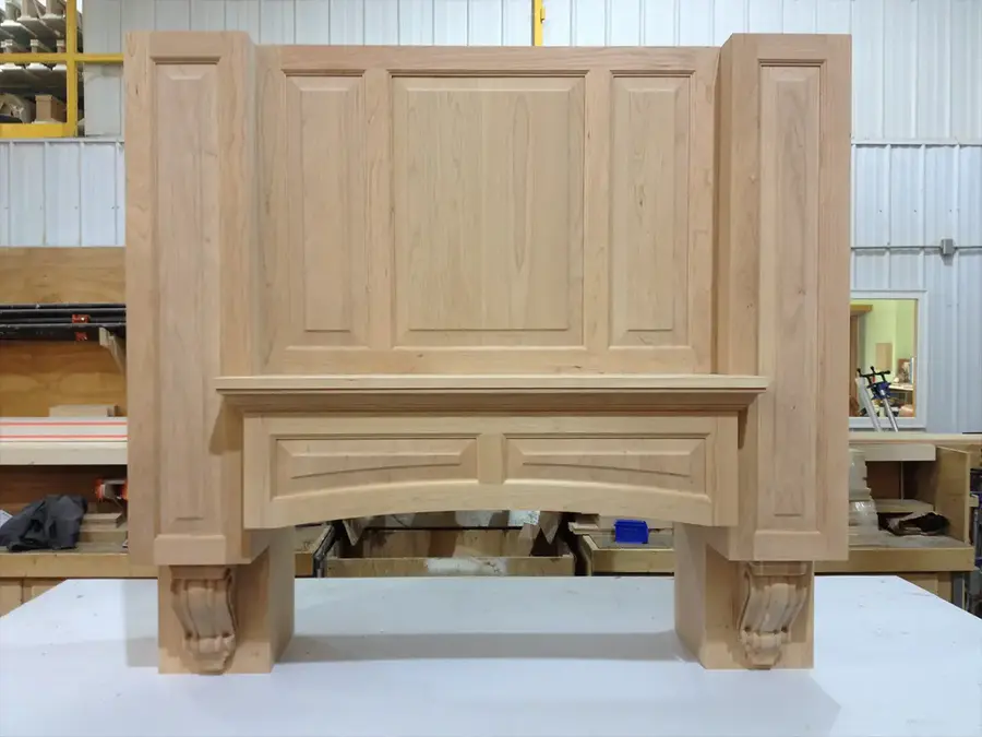 Unfinished raised-panel wood range hood with carved corbels in a millwork shop