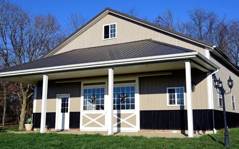 Tan pole barn home with dark metal roof, covered porch and white barn-style doors
