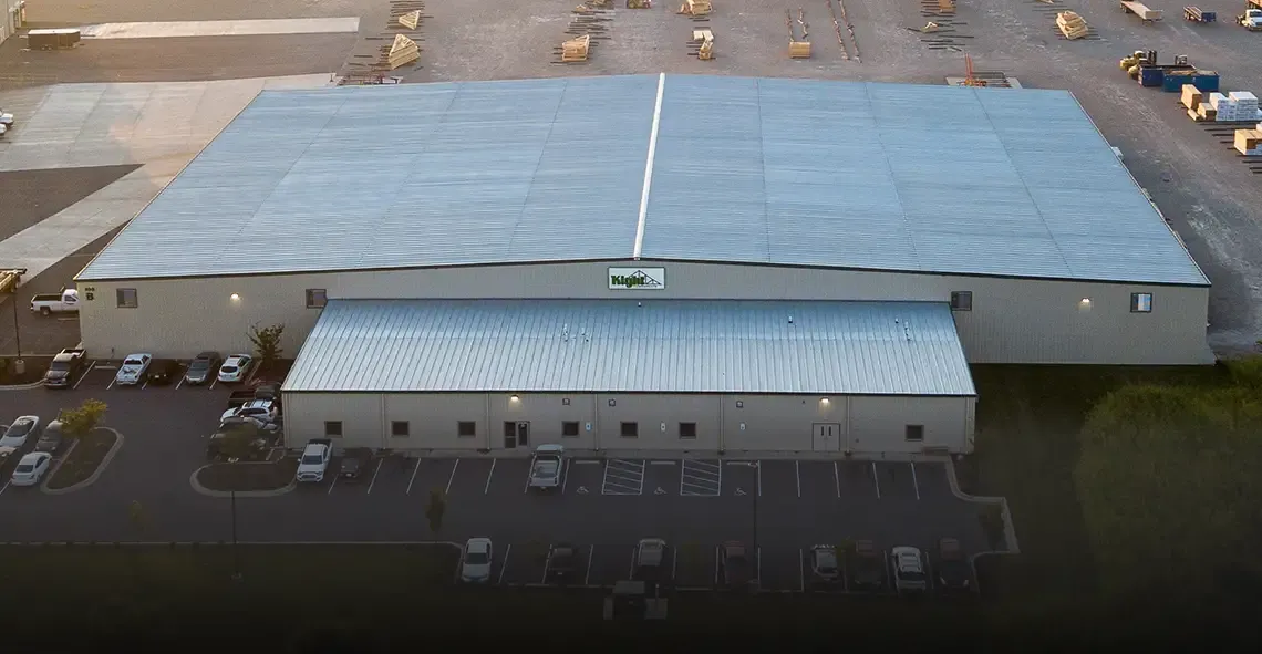 Aerial view of a large metal-roofed component plant with parking lot and lumber yard behind