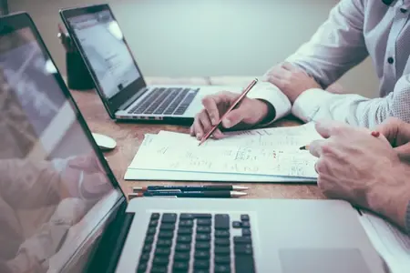 Two people reviewing paperwork at a desk with laptops