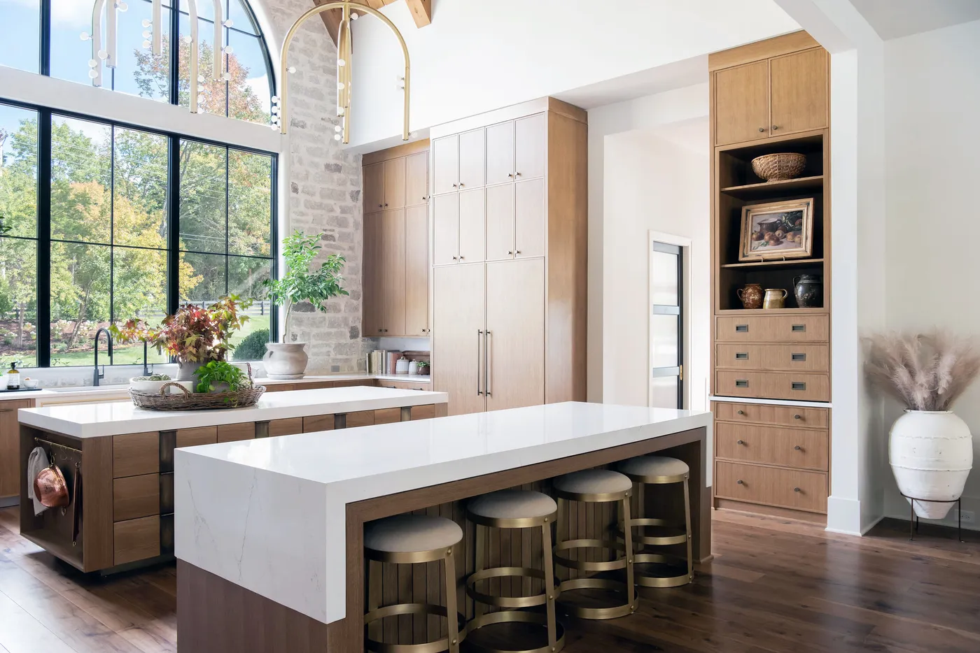 Light wood kitchen with two islands, brass barstools, tall arched windows and stone wall