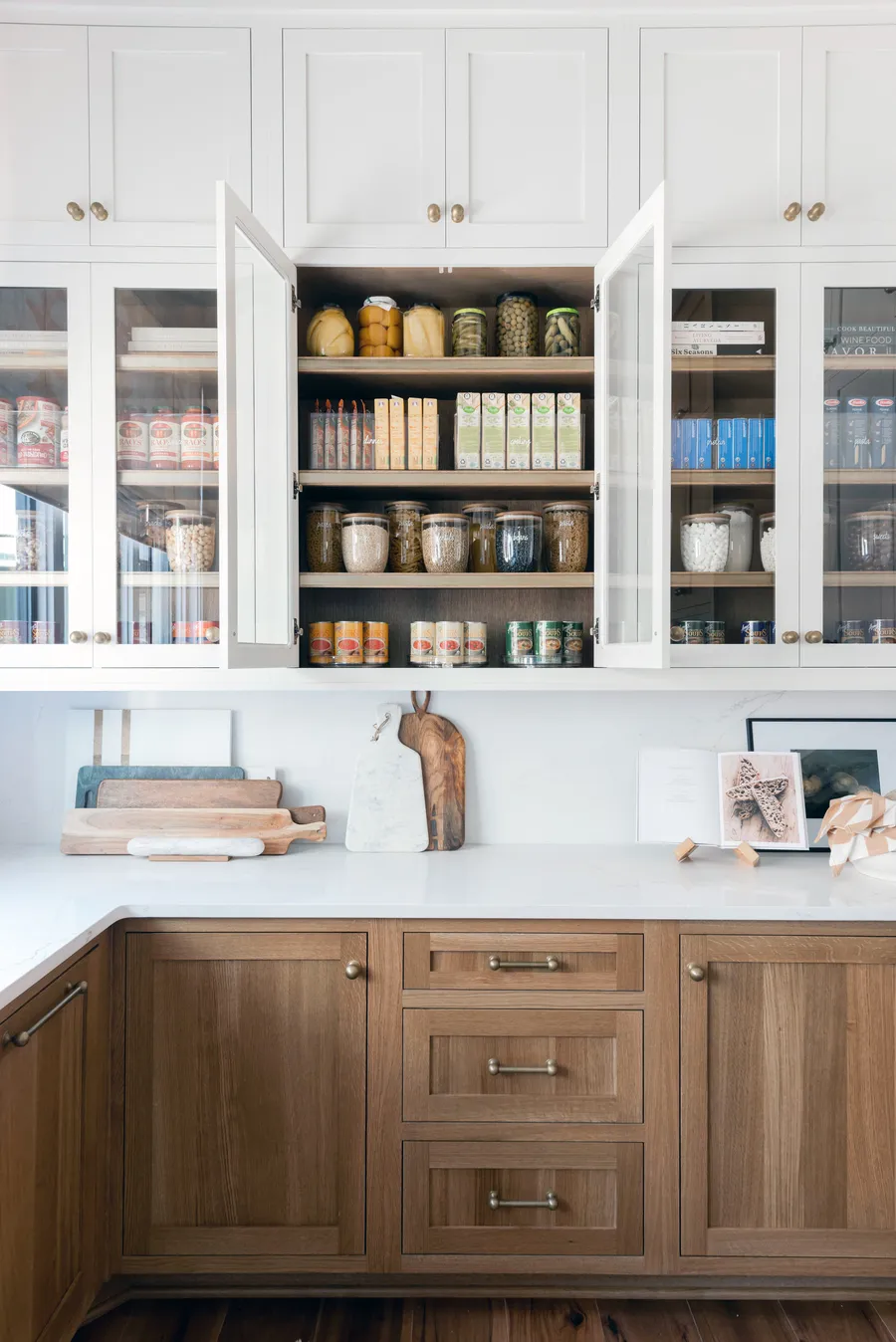 White glass-front pantry cabinets with stocked shelves above light wood base cabinets
