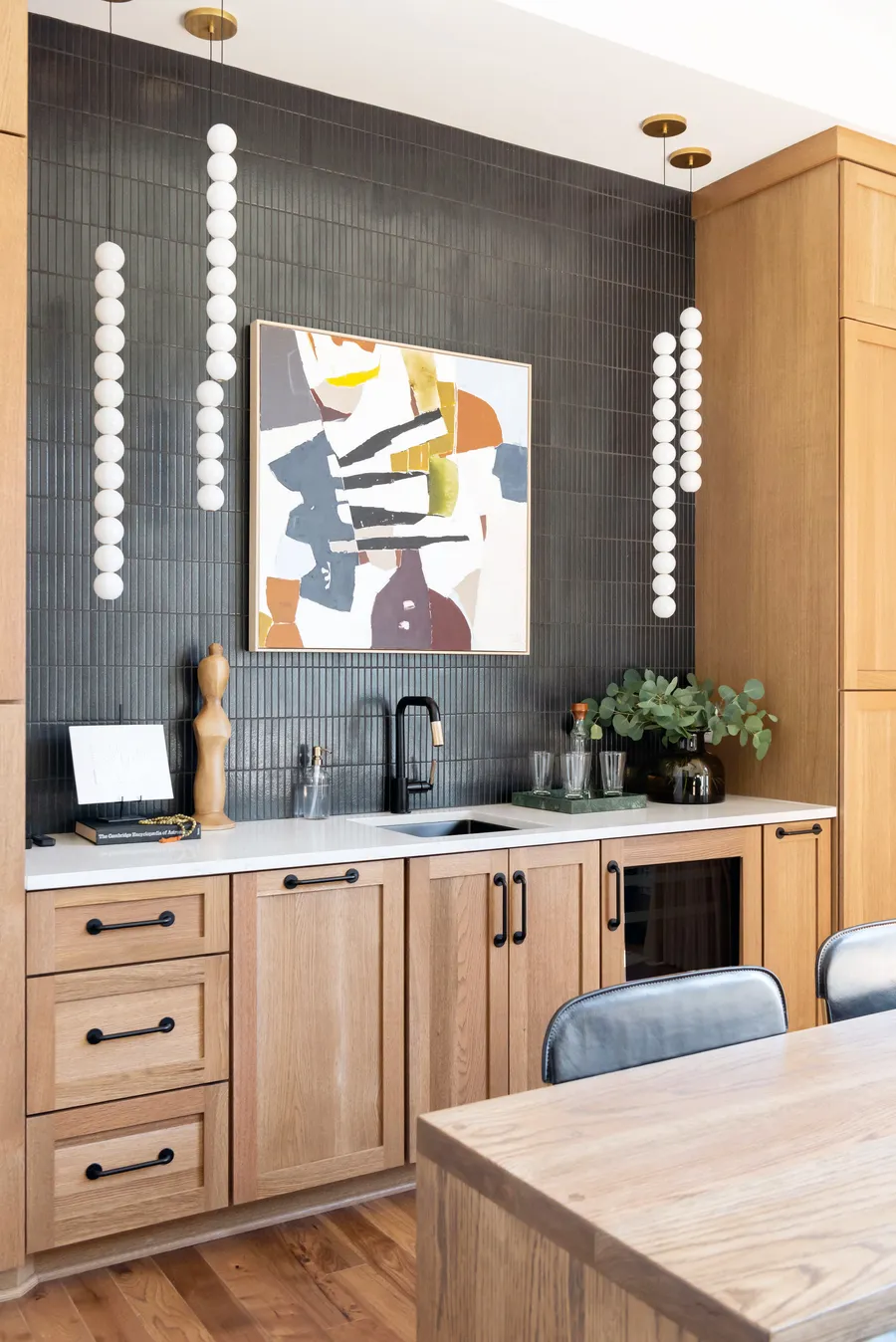 Wet bar with light wood cabinets, black stacked tile, white bead pendants and artwork