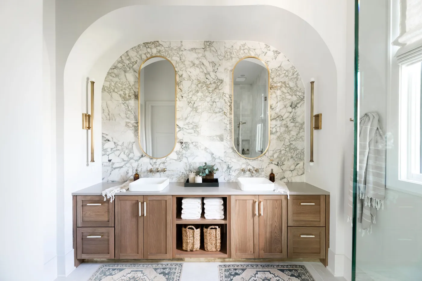 Double vanity with vessel sinks and oval brass mirrors in an arched veined stone niche