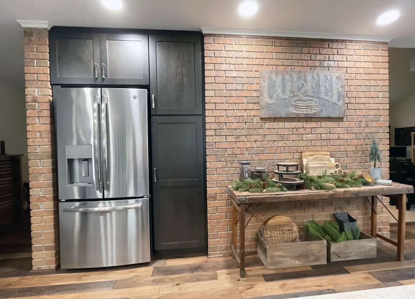 Stainless refrigerator flanked by dark cabinets beside a brick wall coffee station