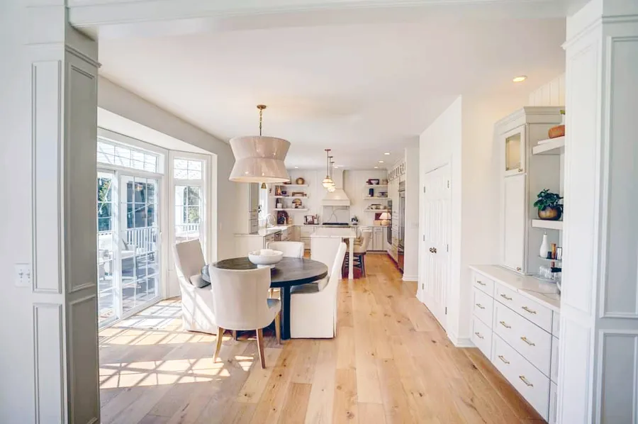 Sunlit dining area with round table and upholstered chairs leading to a white kitchen