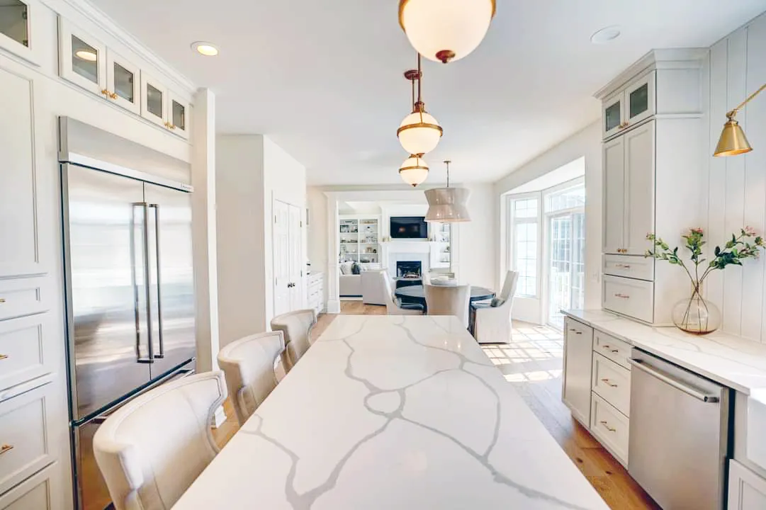Long marble-look island with upholstered stools in a white kitchen opening to living room