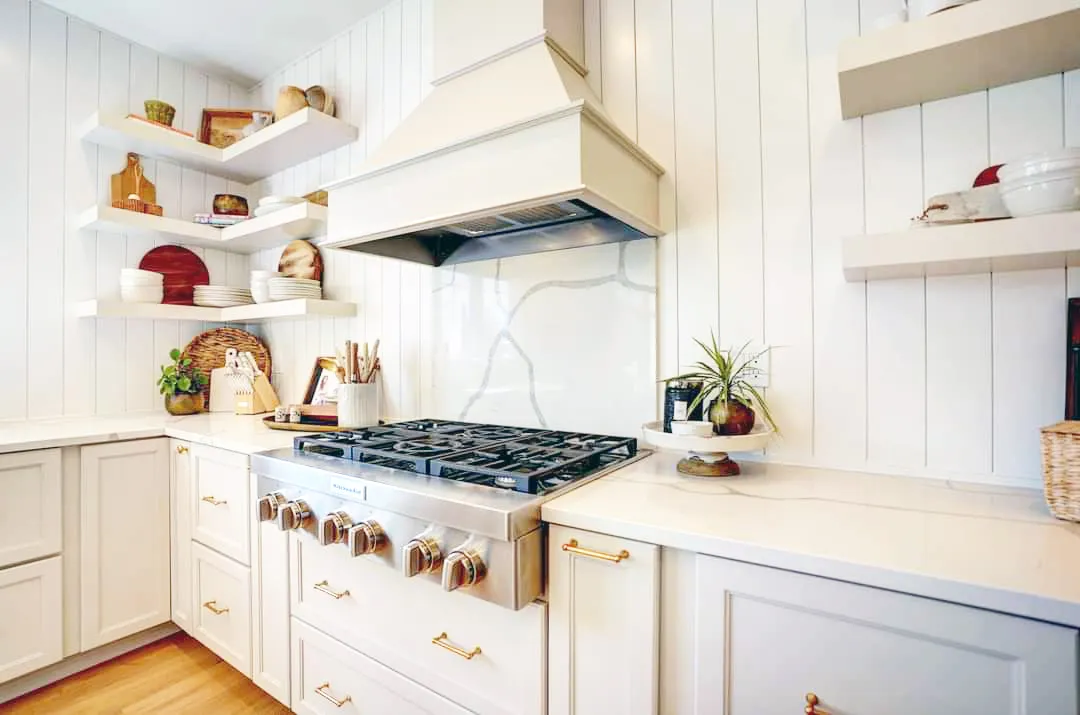 Gas rangetop under a white hood with marble backsplash and open shelves on shiplap